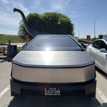 A Tesla Cybertruck parked at a charging stop with a dinosaur statue behind it