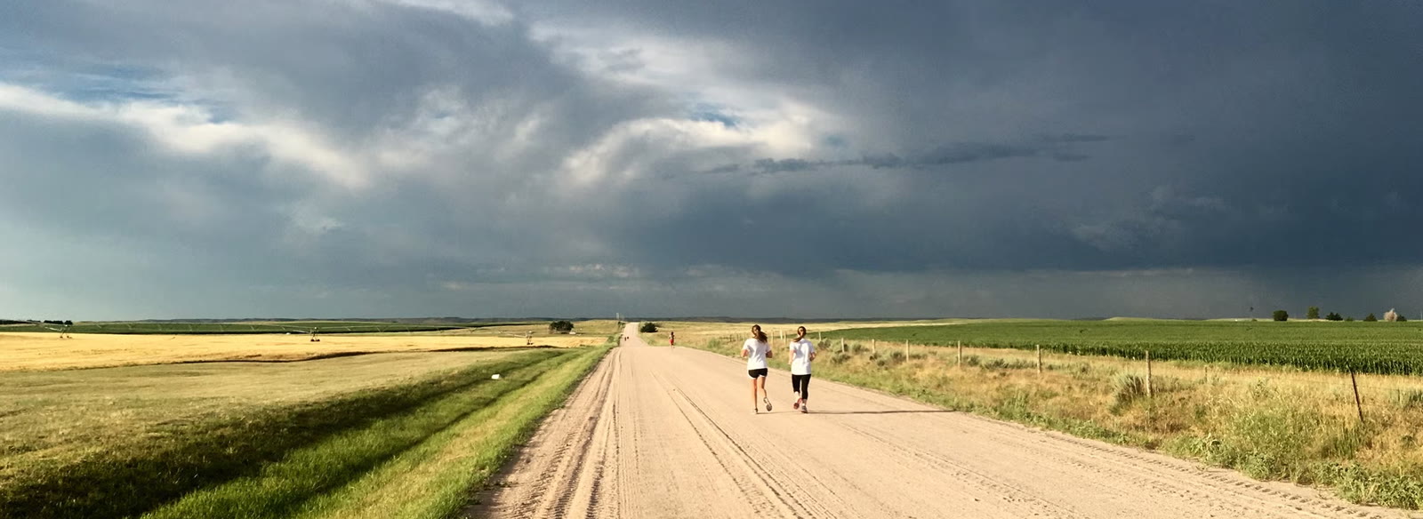 Two runners on a long dirt road under a wide storm sky