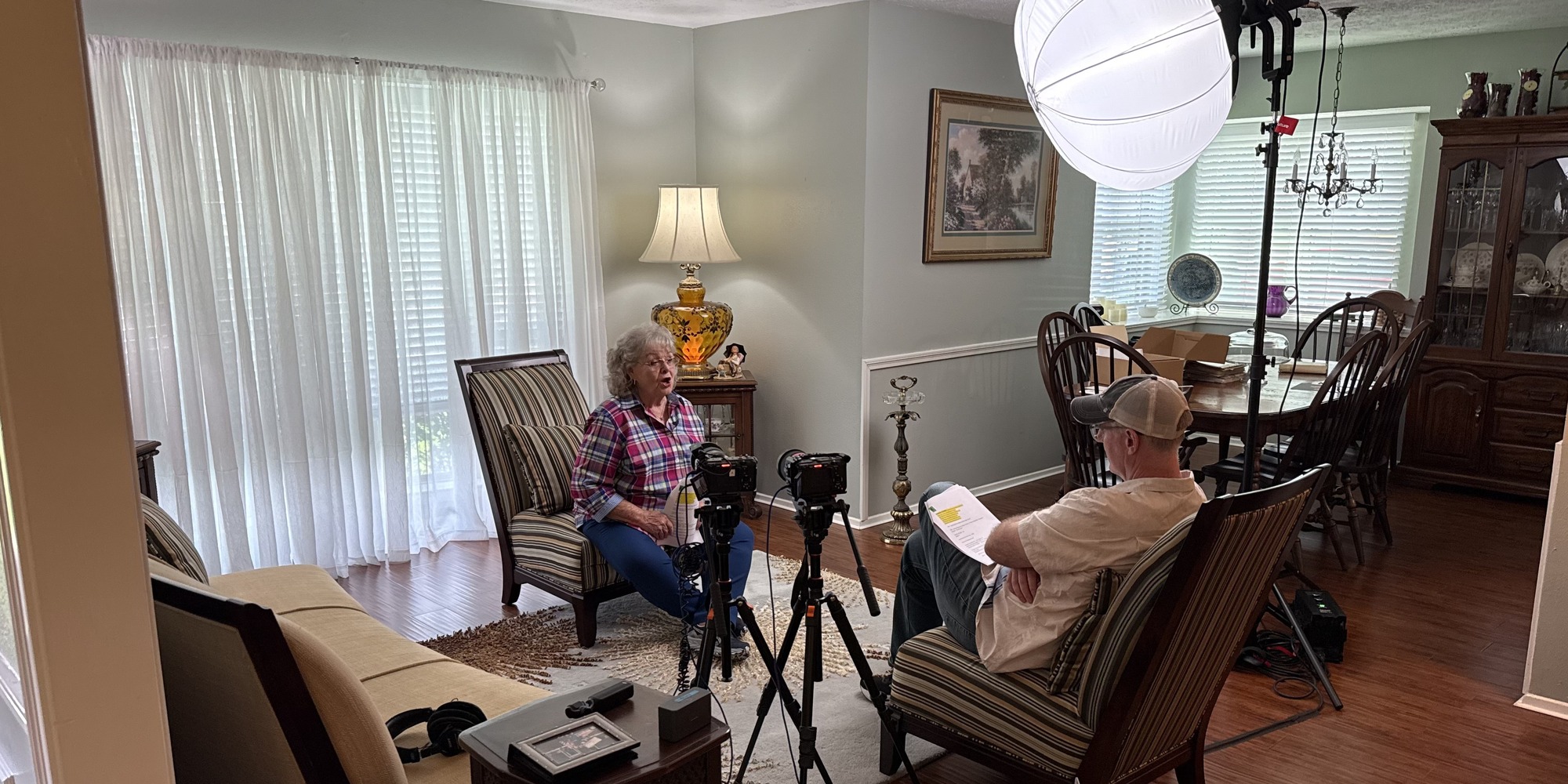 A Heritage Films interview in a living room with two cameras, a softbox light, and an older woman speaking to the interviewer